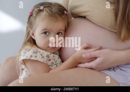 A pregnant Caucasian mother and her daughter are indoors in a living room. The daughter is touching her mother's stomach Stock Photo