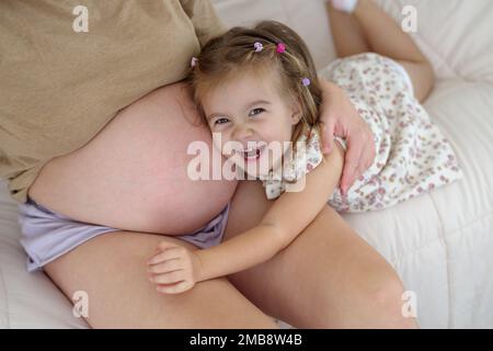 A pregnant Caucasian mother and her daughter are indoors in a living room. The daughter is touching her mother's stomach Stock Photo