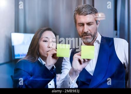 Serious adult man and woman in formal outfits standing near office window and writing ideas on yellow sticky notes Stock Photo