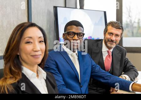 Group of positive diverse coworkers in formal clothes sitting together at table and looking at camera in modern office Stock Photo