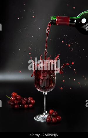 Red wine is poured into a wine glass on a black background with grapes and splash Stock Photo
