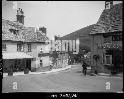 Corfe Castle, Purbeck, Dorset, 1927. A view from The Square showing The Greyhound Hotel on the left and The Bankes Arms Hotel on the right with a man walking past. Both pubs are displaying signs advertising the brewery Strong &amp; Co of Romsey Ltd. Stock Photo