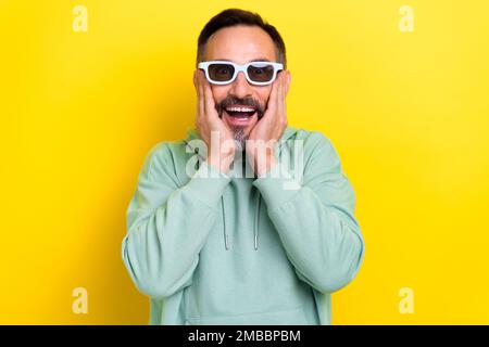 Photo of mature age funny reaction positive man hobby watch films wear glasses touch cheeks excited moment isolated on yellow color background Stock Photo