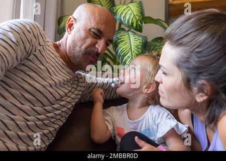 daddy makes funny faces while his little daughter eats baby food with a spoon and mom loves everyone so much, little family gathered in the living roo Stock Photo
