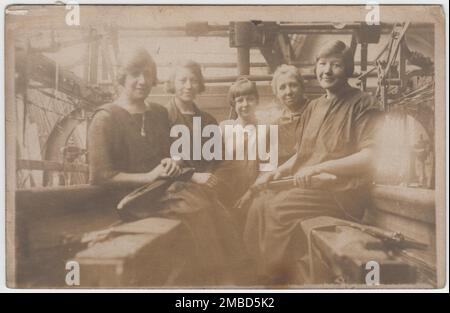 Early 20th century photograph of five women mill workers shown sitting on benches in the factory in between rows of cotton looms. Three generations of women are included in the picture. Two of the women are holding weaving power loom shuttles