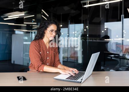 Serious young hispanic woman working in the office on a laptop. Concentrated typing on the keyboard, copying. Stock Photo