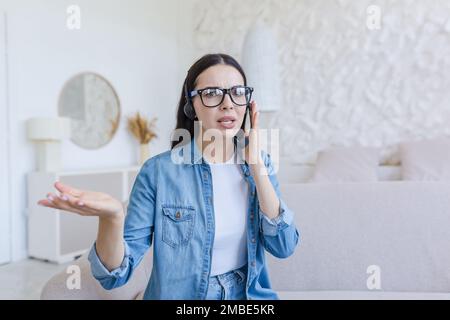 Worried young woman sitting at home on sofa in headset, talking through microphone, online consultation. She looks into the camera, explains, gestures with his hands. Stock Photo