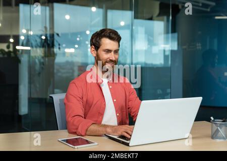 Successful businessman in red shirt happily working with laptop inside office, mature man with beard at workplace typing on keyboard smiling satisfied with work results and achievement. Stock Photo