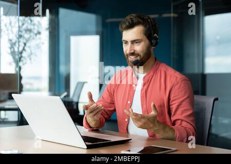 Mature successful businessman with video call headset working inside office, man consulting online clients, talking with colleagues on online remote meeting, sitting at workplace with laptop. Stock Photo