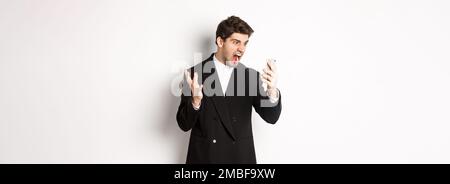 Portrait of angry businessman in black suit yelling at mobile phone, having an argument on video call, standing mad over white background Stock Photo