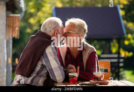 Senior couple sitting on terrace in backyard, drinking coffee and kissing Stock Photo