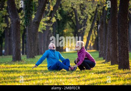 Mature man sitting on grass in park after injury while exercising with wife in nature. Pain in ankle Stock Photo