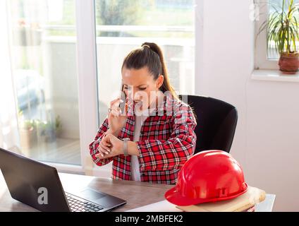 Angry woman construction engineer or architect talking on phone and looking at wrist watch at office Stock Photo