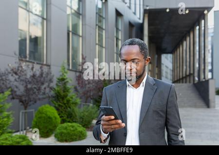 Serious afro american boss in business suit walking outside office building, mature man holding phone, businessman concentrating reading message and typing text. Stock Photo