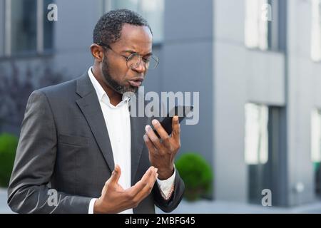 Angry and angry boss talking and shouting on mobile phone, african american businessman in business suit walking outside office and talking with subordinates. Stock Photo