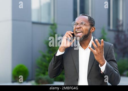 Angry and angry boss talking and shouting on mobile phone, african american businessman in business suit walking outside office and talking with subordinates. Stock Photo