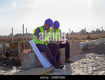 Two engineers with helmets and vests looking at tablet and talking about project at building site Stock Photo