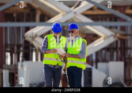 Two engineers with helmets and vests talking at building site in front of metal construction Stock Photo