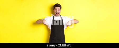 Excited waiter in black apron pointing fingers down, checking out promo offer, standing over yellow background Stock Photo