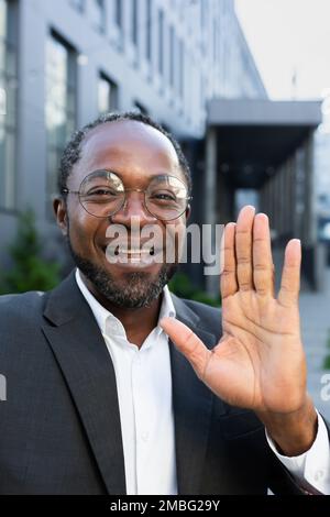Vertical shot, video call of successful businessman boss, African American man outside office building looking into smartphone camera smiling and having fun talking to colleagues online remotely Stock Photo