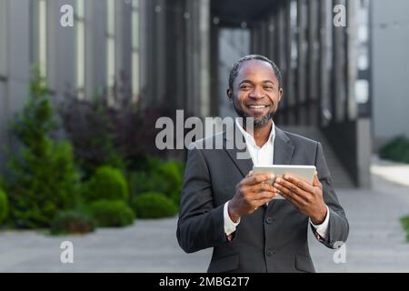 Portrait of a successful African American boss, a businessman in a suit smiling and looking at the camera, a man holding a tablet computer in his hands, a successful investor browsing the Internet. Stock Photo