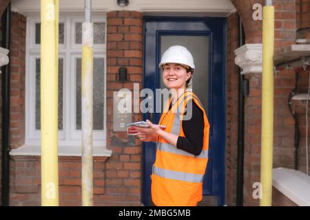 Side view of a smiley civil engineer lady working supervising repair works in a construction site with scaffolding, wearing hard hat orange high vis Stock Photo