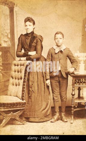 Unidentified standing figures: woman, her folded arms leaning on back of chair, and young man, probably her son, left hand on book, 1890. Stock Photo