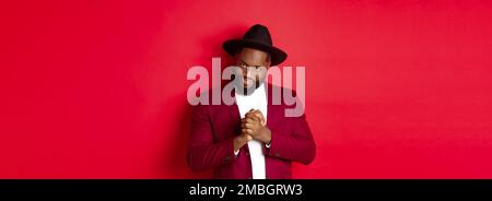Cunning black man having an idea, smiling devious and rubbing hands, standing against red background and scheming Stock Photo