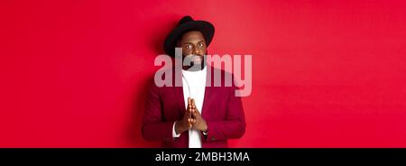Serious looking man getting ready to business, holding hands pressed together and nervously staring at camera, listening closely, standing against red Stock Photo