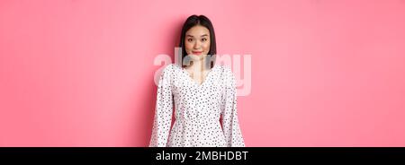 Beautiful asian lady smiling at camera, wearing cute romantic dress, standing over pink background