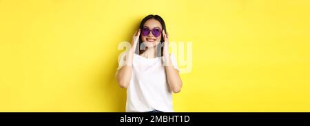 Fashion and lifestyle concept. Stylish asian woman trying new sunglasses in heart-shape, going on vacation, smiling happy at camera, standing over Stock Photo