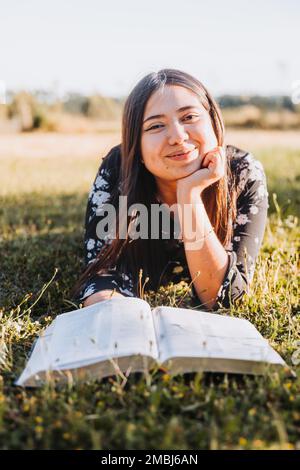 Smiling christian girl lying on the grass reading her bible, in the field at sunset. Looking at camera Stock Photo