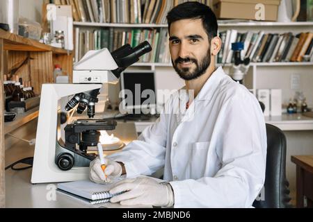 Young scientist in front of microscope, smiles and looks at camera and takes note of the samples he is examining in the lab. Stock Photo