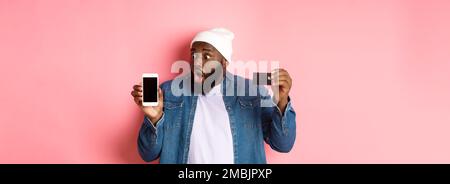Online shopping. Impressed and surprised Black man showing credit card, staring at smartphone screen, demonstrate app, standing over pink background Stock Photo
