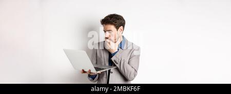 Shocked office worker in suit looking worried at laptop screen, having trouble at work, standing on white background Stock Photo