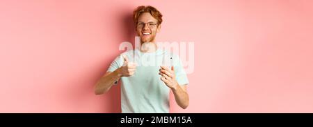 Happy young man with red messy hair, showing thumbs-up while using mobile phone, smiling and praising application, standing over pink background Stock Photo