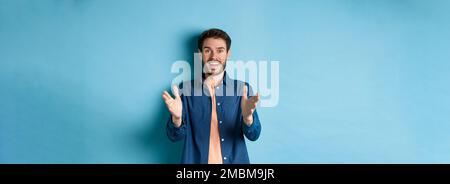 Cheerful young man pointing hands at camera and smiling, feeling glad for you, praising nice work, standing on blue background Stock Photo