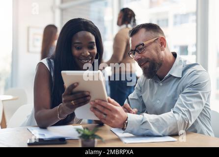 We work better when we work together. two young businesspeople sitting together and discussing in the office while their colleagues work behind them. Stock Photo