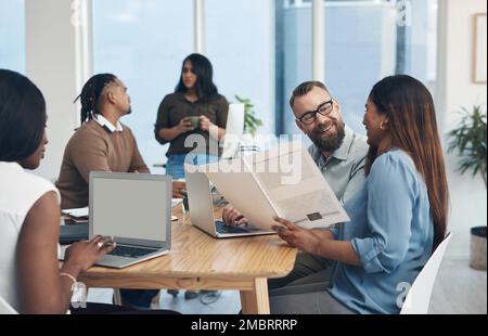 Weve got great ideas. two young businesspeople sitting together and reading through paperwork while their colleagues work in the background. Stock Photo