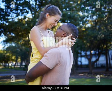Love, hug and interracial couple at a park, happy and excited against a tree background. Romance, sweet and black man lifting his girlfriend, in love Stock Photo