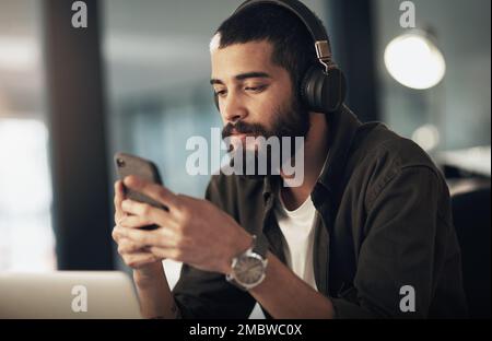 Get powered up with a good playlist. a young businessman using a smartphone with headphones during a late night at work. Stock Photo