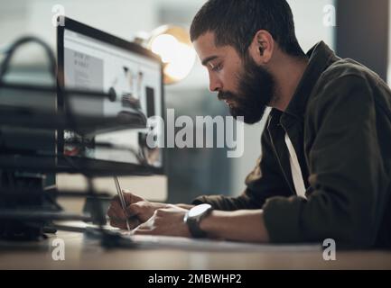 Working his way to employee of the month. a young businessman writing in a notebook and using a computer during a late night at work. Stock Photo