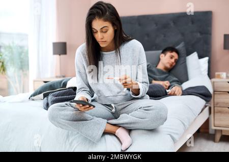 If you dont know, Doctor Internet might. a young woman using a smartphone and thermometer while recovering from an illness with her husband at home. Stock Photo