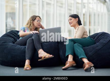 Using their proven strategies to gain the best advantages. two businesswomen using a laptop together while sitting on beanbags in an office. Stock Photo