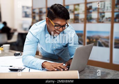 Connecting his designs with the world. a young architect using a laptop while drawing up a building plan. Stock Photo