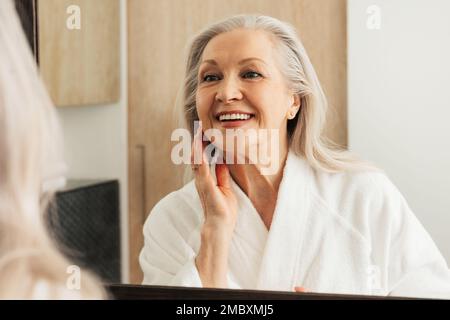 Happy aged female touching her face in front of a mirror. Woman in bathrobe admires her reflection. Stock Photo