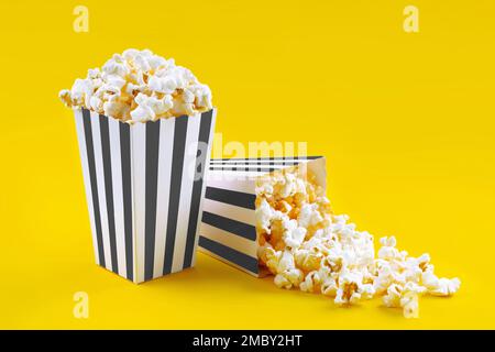 Two black white striped carton buckets with tasty cheese popcorn, isolated on yellow background. Box with scattering of popcorn grains. Movies, cinema Stock Photo