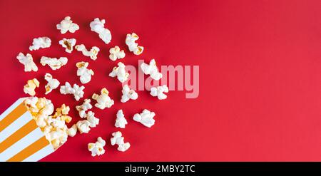 Tasty cheese popcorn falling out of a orange striped carton bucket, isolated on red background. Scattering of popcorn grains. Movies, cinema, fast foo Stock Photo