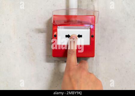 Close up of male hand pointing at red fire alarm switch on concrete wall in office building. Industrial fire warning system equipment for emergency. Stock Photo