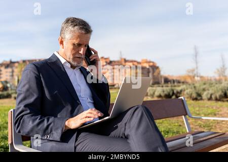 Concentrated senior businessman in formal wear sitting on bench and talking via smartphone while browsing laptop during remote work Stock Photo